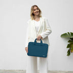 Woman in a white blazer and wide-leg trousers standing against a white wall holding a teal MOSISO laptop bag by its top handles with a tropical plant in the background