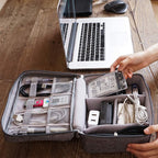 Person organizing electronics and cables into an OrgaWise three-layer travel organizer on a wooden desk next to laptop