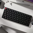 Top-down view of Keychron K2 V3 75% mechanical keyboard on a white desk with red Escape keycap and subtle RGB underglow, alongside open AirPods case, iMac base, and scroll wheel of a mouse partially visible at edges