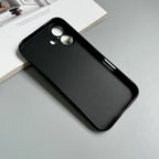 BESDYNASTY matte black silicone iPhone 16 case lying flat on gray surface leaning against open white book, back view showing smooth matte TPU finish and dual circular camera cutout with microphone hole, slim black TPU frame visible around edges