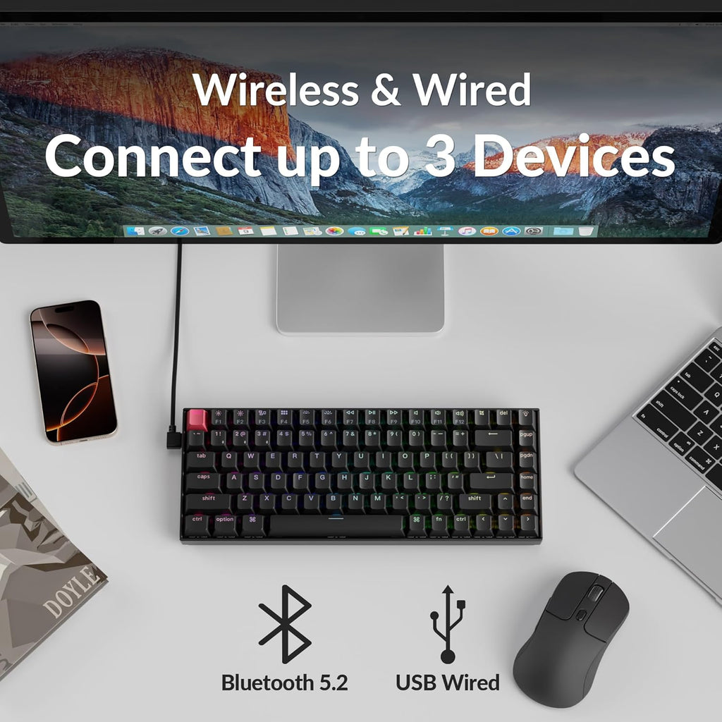 Top-down flat lay of Keychron K2 V3 keyboard on a white desk connected via USB-C cable to an iMac, with a MacBook, iPhone, wireless mouse, and magazine nearby, with Bluetooth 5.2 and USB Wired icons and text reading Wireless and Wired Connect up to 3 Devices