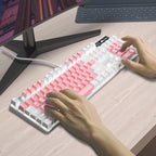 Person using a pink and white keyboard on a wooden desk with a computer monitor in front of them