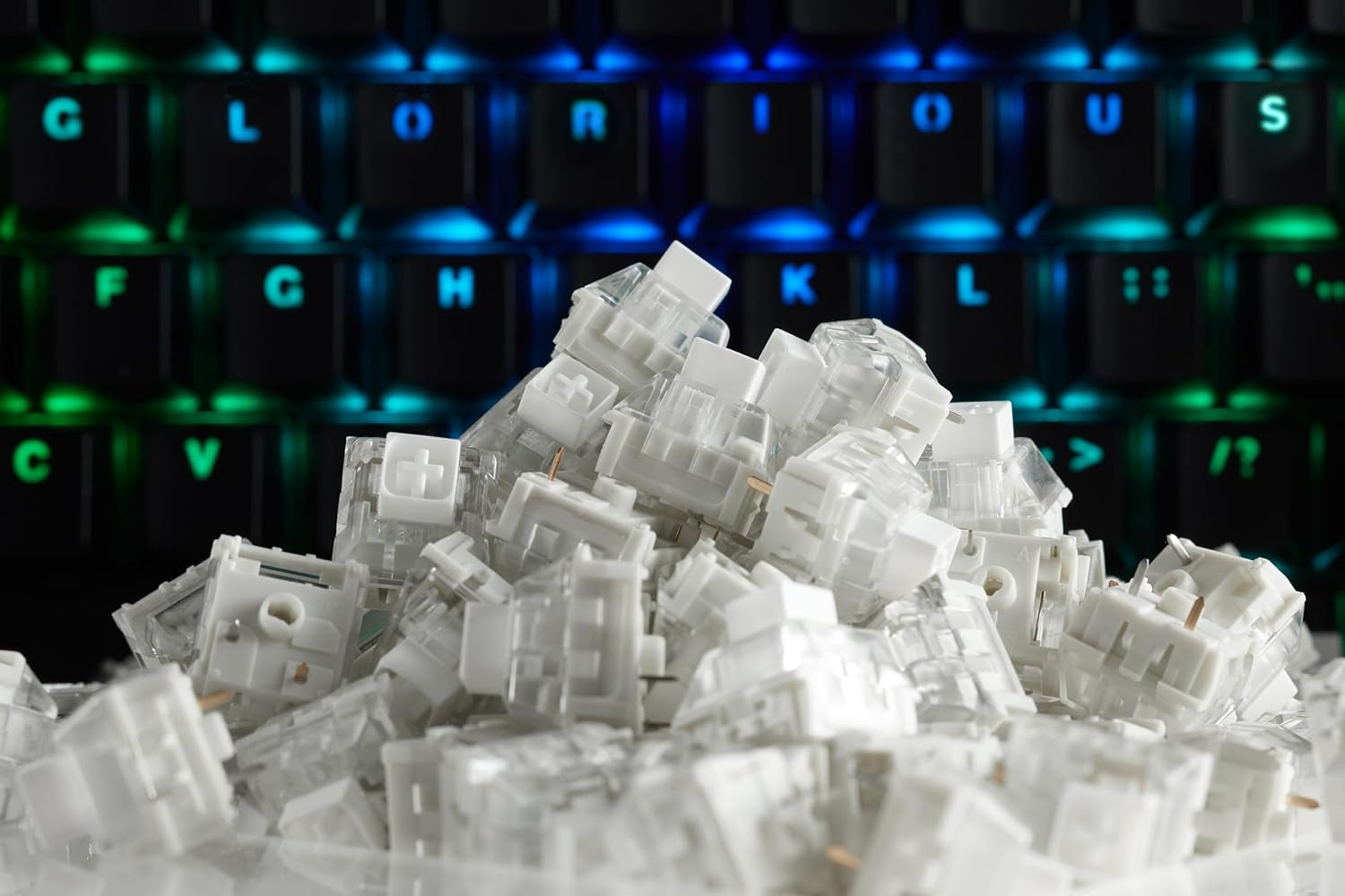 Large pile of white mechanical keyboard switches in front of a Glorious RGB mechanical keyboard with blue and green backlighting