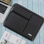 Lacdo black laptop sleeve laid flat on a light blue wooden surface next to an open MacBook Pro and a magazine, showing front pocket layout and silver zipper pulls