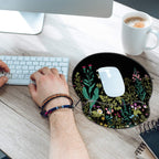 Hand resting on iDonzon garden flowers gel wrist rest mouse pad on a light wood desk with white Apple Magic Mouse on the pad surface, white Apple keyboard to the left, white coffee mug and spiral notebook partially visible, with overhead natural lighting