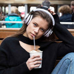Young woman with brown hair wearing white COWIN E7 over-ear headphones outdoors on a wooden bench in a crowded public space, holding a whipped cream topped drink with a straw, wearing a black long-sleeve top and blue jeans
