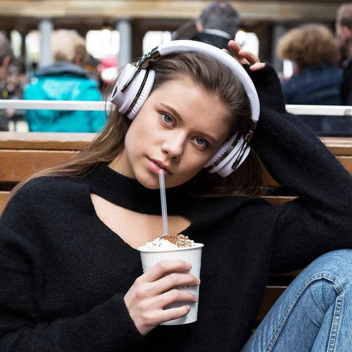 Young woman with brown hair wearing white COWIN E7 over-ear headphones outdoors on a wooden bench in a crowded public space, holding a whipped cream topped drink with a straw, wearing a black long-sleeve top and blue jeans