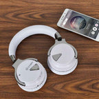 White COWIN E7 headphones laid flat on a dark walnut wood surface next to a white Android smartphone displaying a music app playing Imagine Dragons' Radioactive, showing the outer ear cups, chrome hinges, and COWIN logo