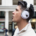 Young man with dark hair wearing white COWIN E7 over-ear headphones outdoors on a city street, shown from a side profile angle looking upward, wearing a cream zip-up sweater with blurred storefronts in the background