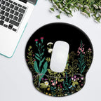 Top-down flat lay of iDonzon round garden flowers gel wrist rest mouse pad on a white surface with white Apple Magic Mouse centered on the pad, silver laptop keyboard visible in upper left corner, and bouquet of white carnations in upper right corner